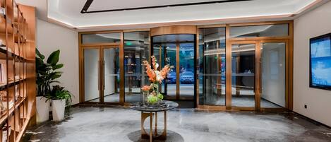 A lobby with a marble floor, a central table with a floral arrangement, and a glass door entrance.