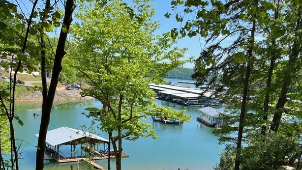 Une terrasse en bois qui mène à un quai surplombant un lac paisible, le tout entouré d’arbres et d’une zone herbeuse.