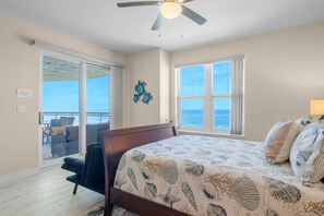 Une chambre avec un lit, un ventilateur de plafond, une vue sur l’océan par la fenêtre et un balcon aménagé avec du mobilier d’extérieur.