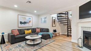 A living room with a grey sofa, a coffee table, and a patterned rug. There is a fireplace, a TV mounted on the wall, and a staircase leading to another level. - New! Refreshing Beach Getaway- Heart of San Diego! (San Diego)