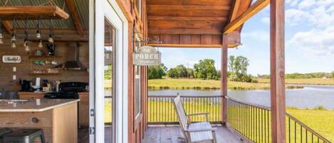 A wooden porch with a sign reading "The Porch," a bar area with stools, and a view of a lake.