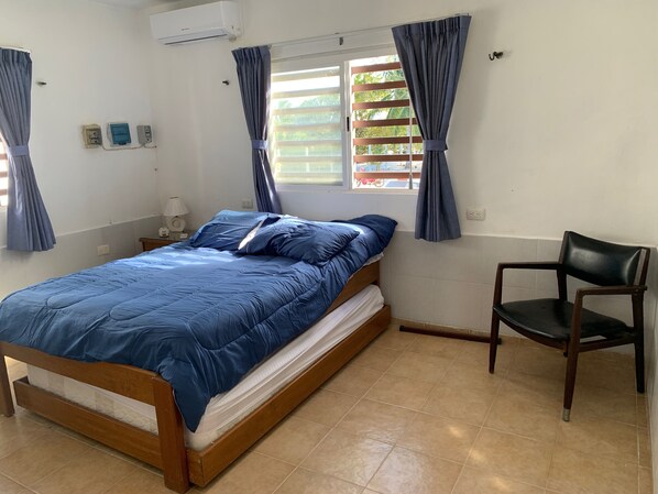 A bedroom with a bed, a chair, a window with curtains, an air conditioner, and a wall socket.