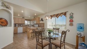 A spacious kitchen with wooden cabinets, a dining area with a glass table and metal chairs, and a large window with blinds. - Mohave desert retreat  (Fort Mohave)