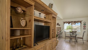 A wooden entertainment center with a flat-screen TV, shelves with decorative items, and a dining area with a table and chairs. - Mohave desert retreat  (Fort Mohave)