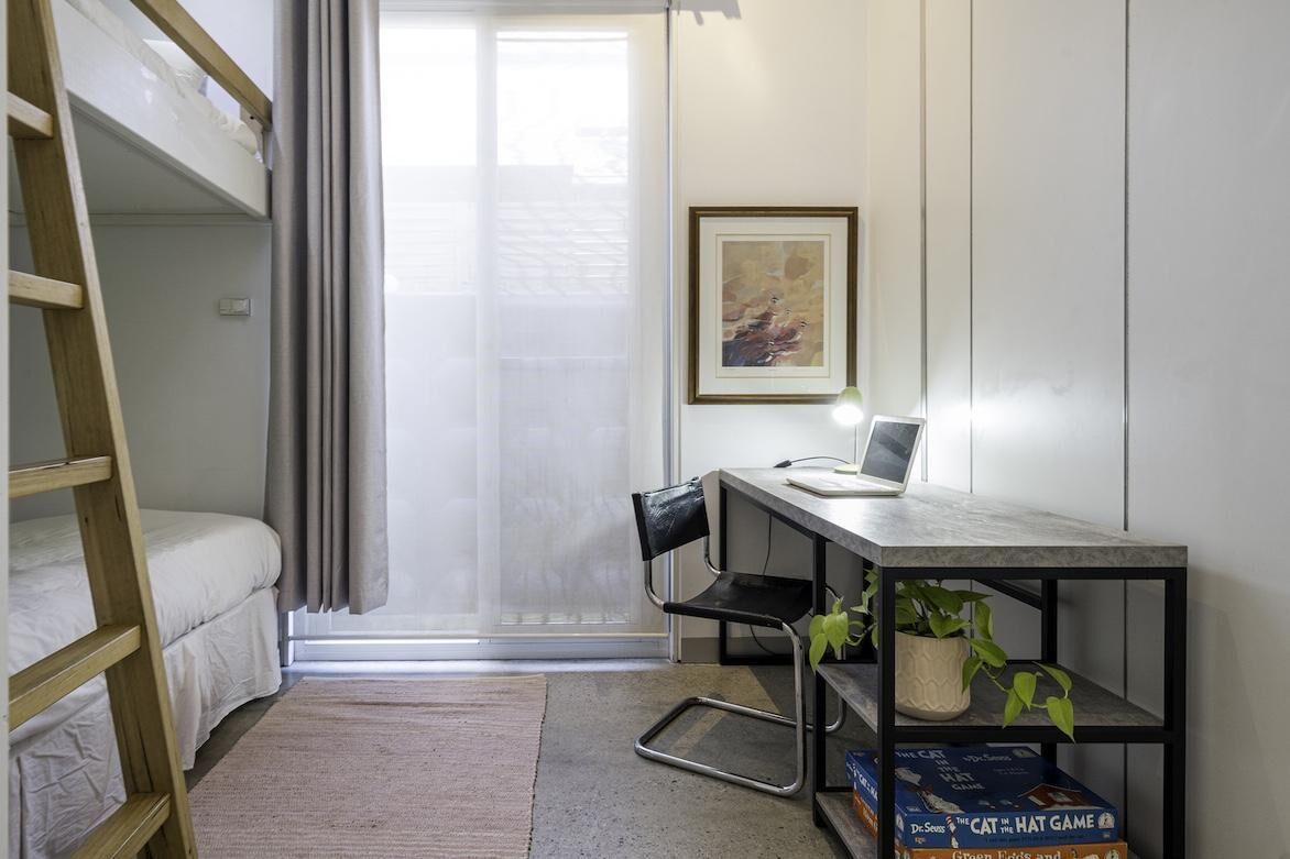 A dorm-style room with bunk beds, a desk with a laptop, a chair, a potted plant, and a framed picture on the wall.