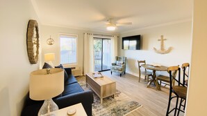 Ein Wohnzimmer mit Sofa, Couchtisch und Lampe. Es gibt einen Essbereich mit Tisch und Stühlen. Ein Deckenventilator ist sichtbar.
