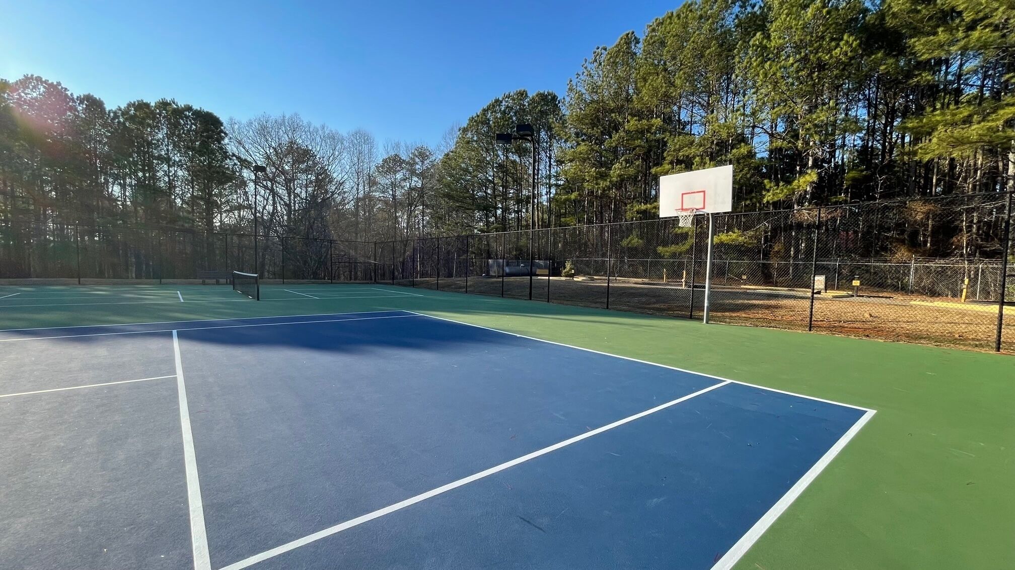 Un terrain de basketball en plein air, doté d’une surface verte et bleue, entouré d’une clôture en treillis métallique et d’arbres.
