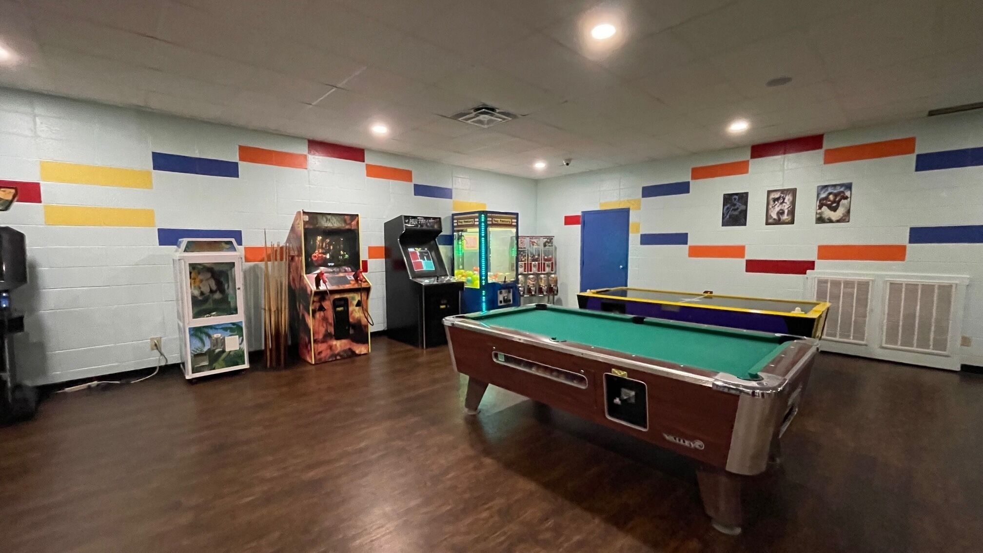A pool table in a room with wooden floors, arcade games, and colorful wall decorations.