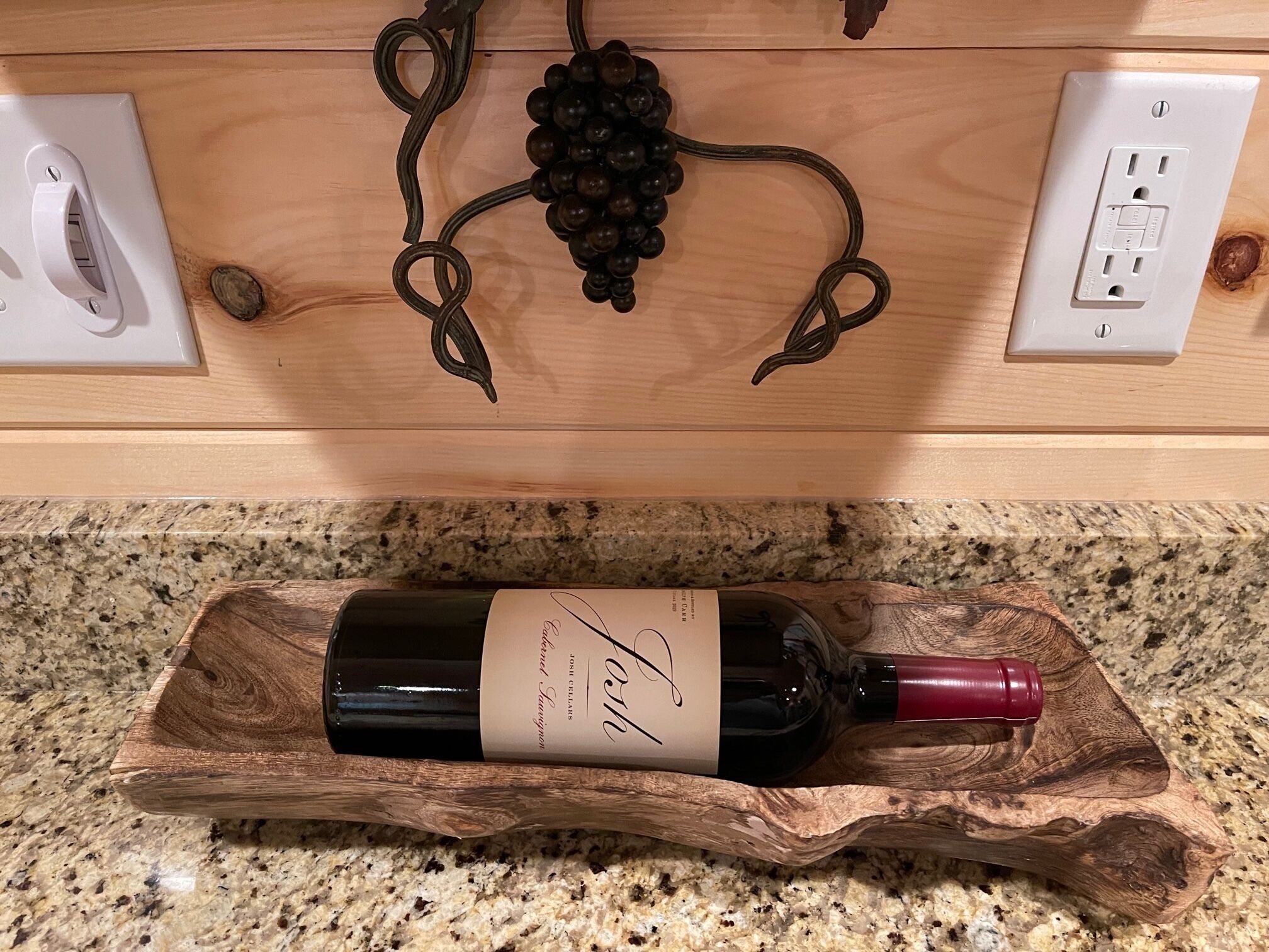 Une bouteille de vin posée sur un plateau en bois dans une cuisine dont le plan de travail est également en bois et qui est équipée de prises électriques.