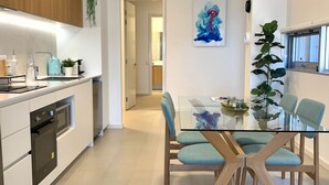 A modern kitchen with built-in appliances, a dining area with a glass table and blue chairs, and a hallway leading to another room. - Mariners Cove Mindarie ~ Luxe Marina Waterfront (Mindarie)