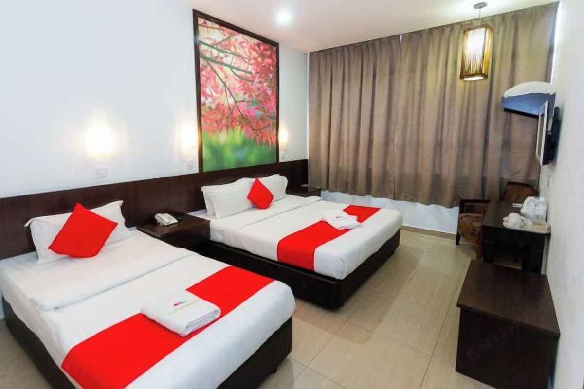 A hotel room with two beds, each with white bedding and red accents, a painting of flowers on the wall, a desk with a lamp, and a television.