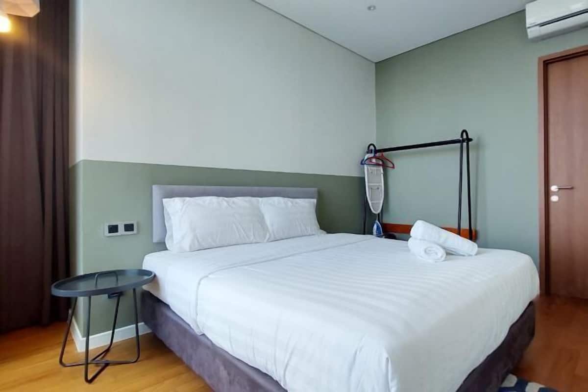 Een netjes opgemaakt bed met witte lakens, een klein rond tafeltje met een stoel en een wandgemonteerde airconditioning.