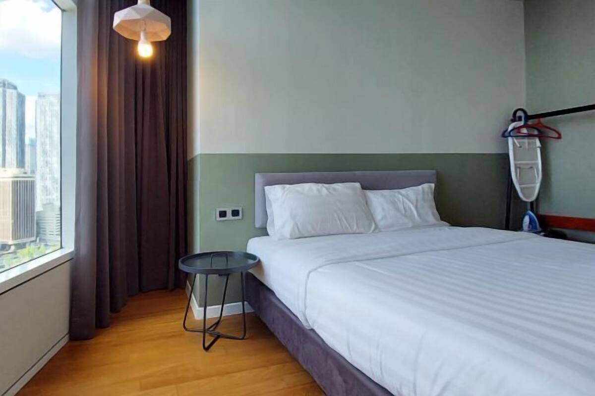 Een hotelkamer met een bed, een kleintje tafeltje, een stoel, een raam met gordijnen en uitzicht op gebouwen.