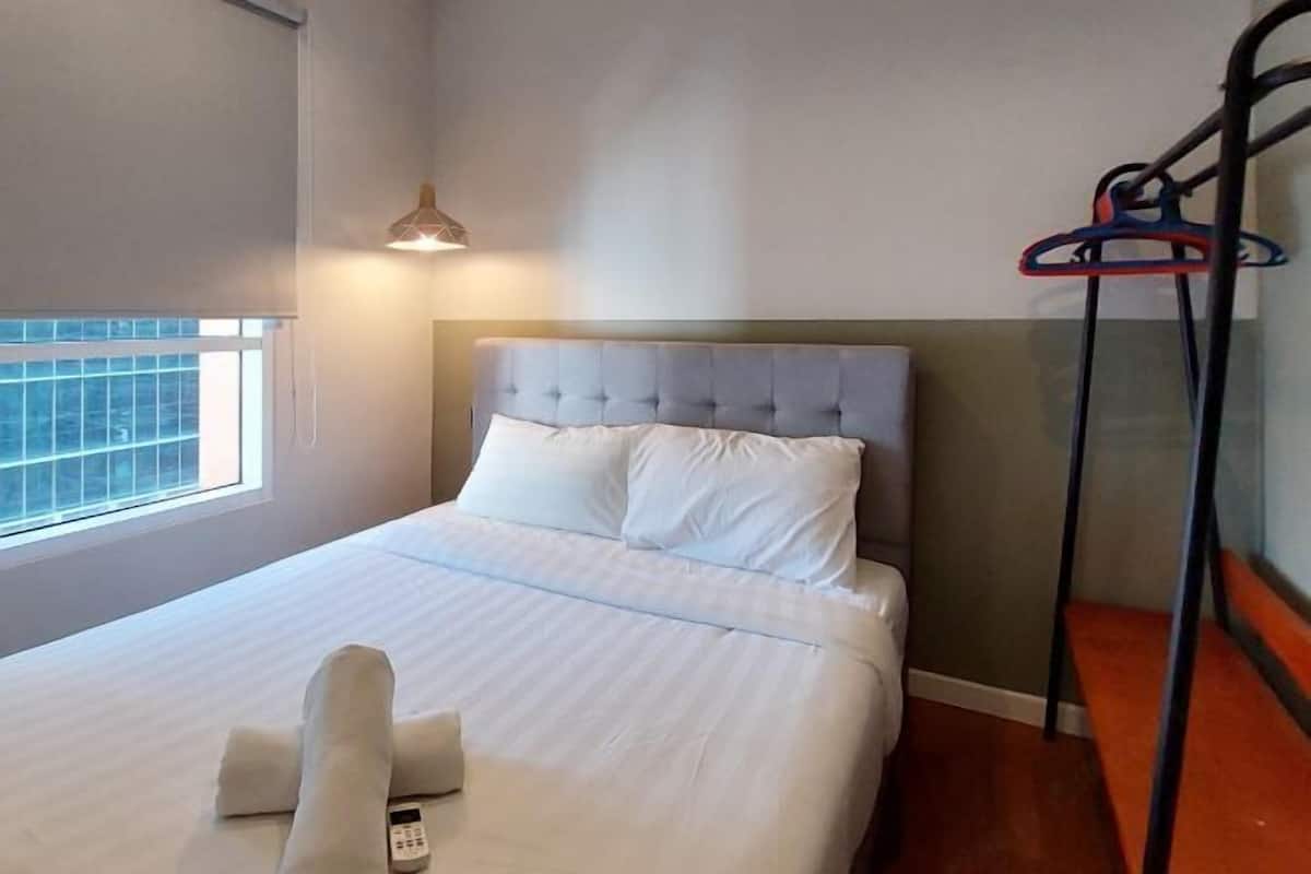 Een net opgemaakt bed met witte lakens en kussens, een nachtkastje met een afstandsbediening, een hanglamp en een raam met uitzicht.