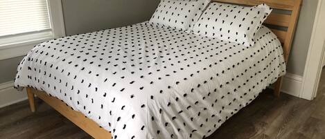 A bed with a white bedspread featuring a black pattern, a wooden headboard, and a wooden floor.