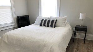 Un lit bien fait, avec une couette blanche, deux coussins à motifs et une petite table de chevet surmontée d’une lampe.