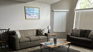 Una sala de estar con dos sofás, una mesa de centro y una silla. Hay una ventana con persianas y un cuadro en la pared.