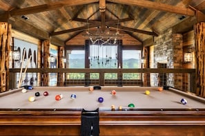 A pool table with balls, wooden beams, and a chandelier in a room with stone walls and a view.