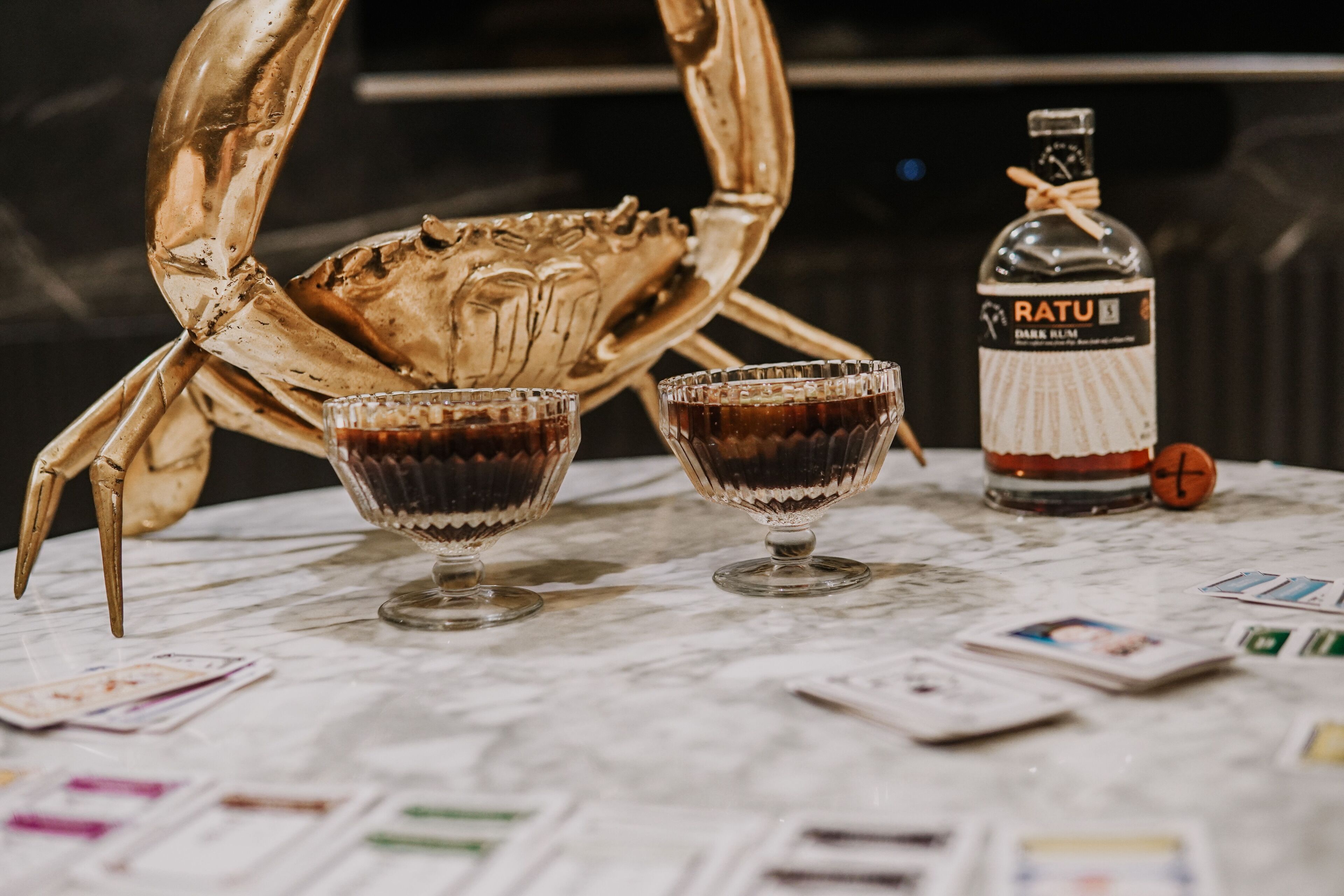 A table with two glasses of dark liquid, a bottle labeled "Ratu," and a golden crab sculpture.