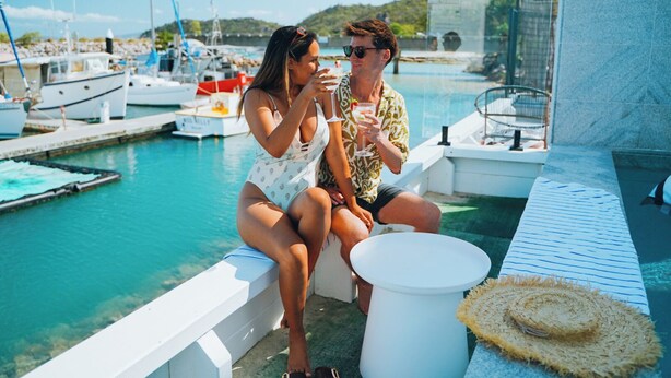 Una pareja sentada junto a una piscina, con barcos al fondo.