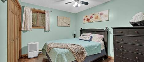 Un dormitorio con una cama, un tocador, un ventilador de techo, una ventana con cortinas y una pared con dos cuadros.