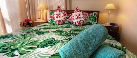 A neatly made bed with a tropical leaf-pattern bedspread, two decorative pillows, and two bedside lamps.
