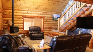 A log cabin interior with a fireplace, leather sofa, and a staircase leading to an upper level. - Peaceful Pines (Custer)