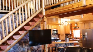 A wooden interior with a staircase, a flat-screen TV, a kitchen area, and a black leather sofa. - Peaceful Pines (Custer)