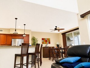 A modern kitchen with wooden cabinets, a dining area with chairs, a black leather sofa, and a ceiling fan.