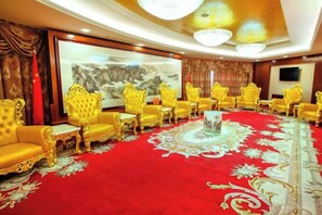 Un salón de conferencias con sillas doradas dispuestas alrededor de una mesa central, una alfombra roja y un cuadro grande en la pared.