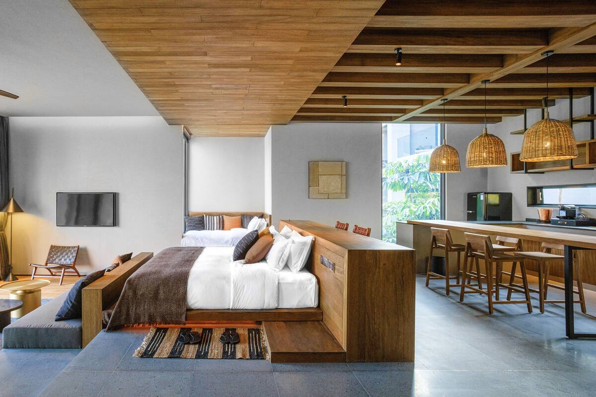 Una habitación de hotel moderna con cama, zona de estar, kitchenette y ventanas amplias.
