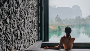 Uma pessoa sentada perto de uma janela, com vista para águas calmas e montanhas.