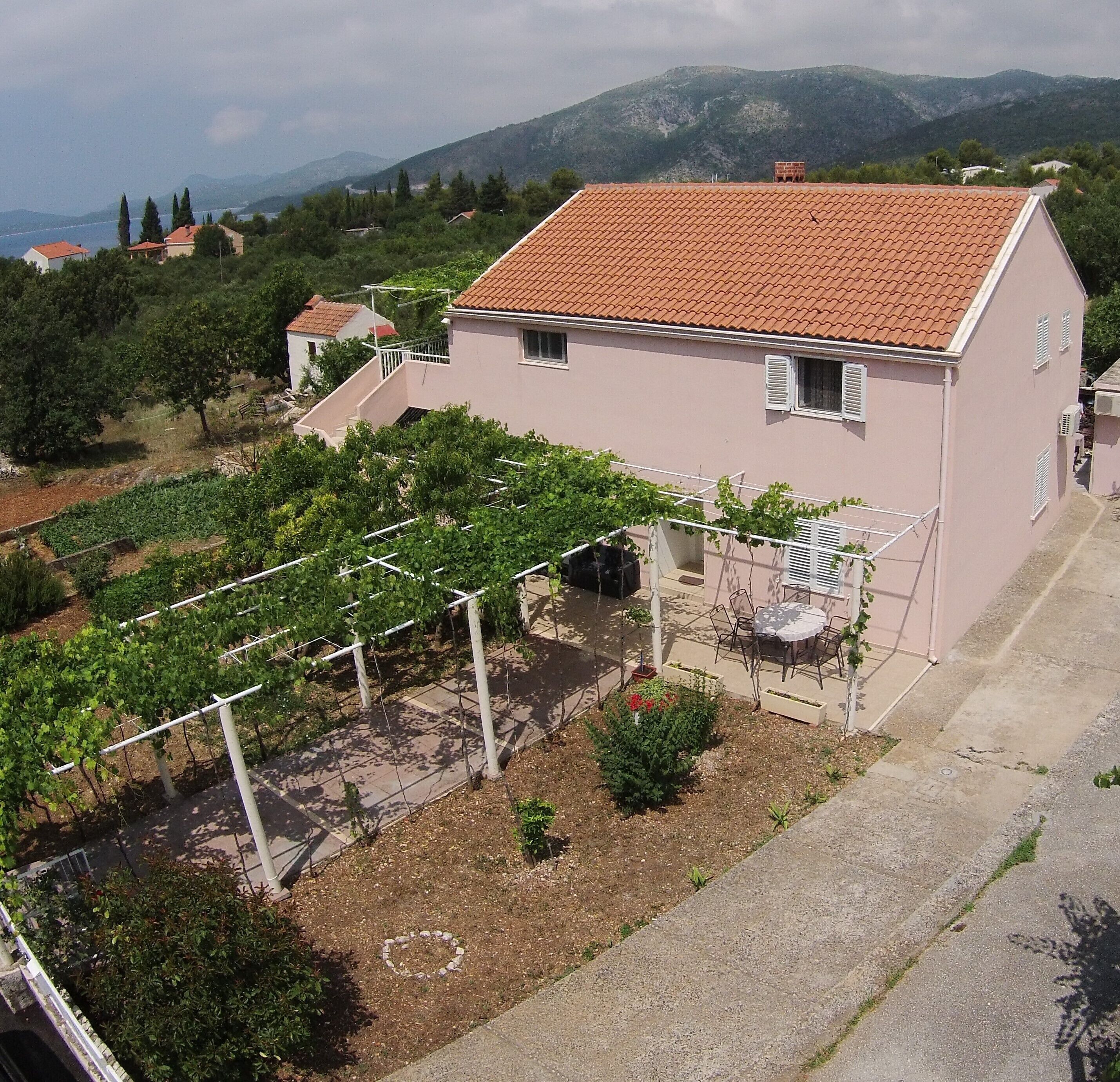 A house with a red-tiled roof, a garden with grapevines, and a view of mountains in the background.