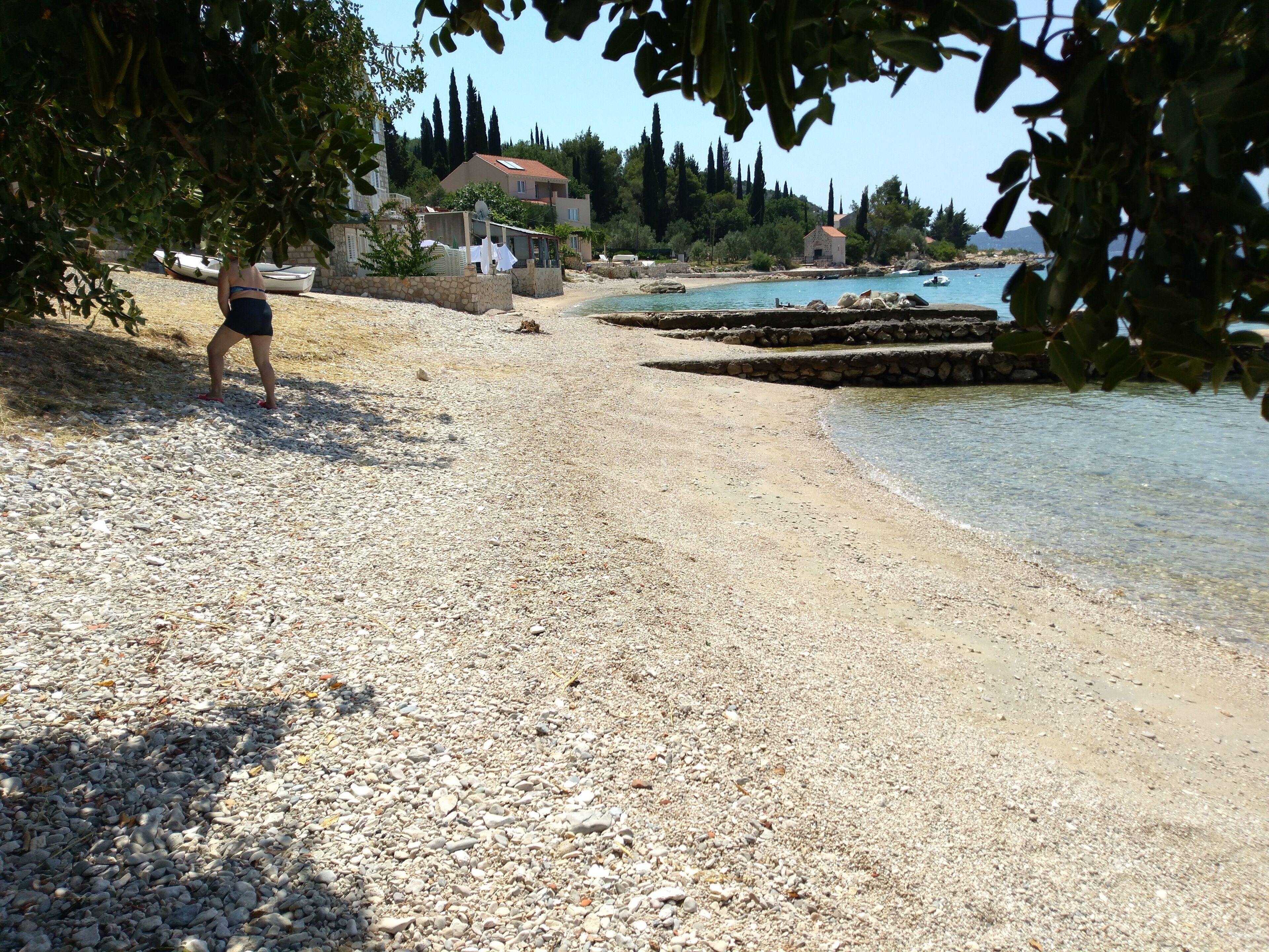 Ein Kiesstrand mit einer Person, die spazieren geht, Häusern im Hintergrund und einem klaren Himmel.