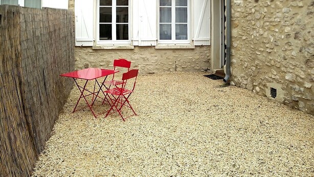 A building with a stone facade, white shutters, and a red table with chairs in the courtyard.