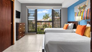 Habitación de hotel con dos camas, televisión, cómoda y balcón con vistas a palmeras y edificios.