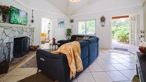 Ein Wohnzimmer mit Kamin aus Stein, einem großen Fernseher, einem blauen Sofa und Blick auf den Garten durch eine Glastür.