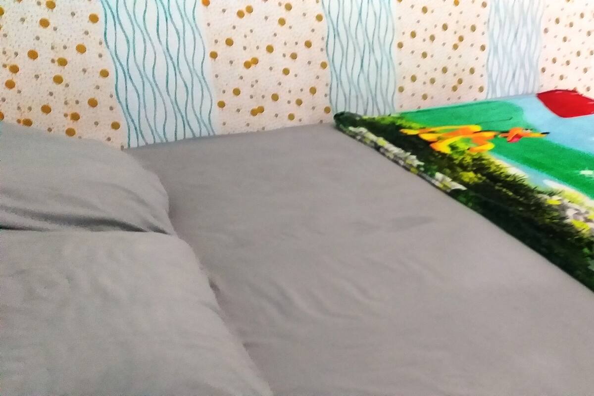 Un lit avec des draps gris, une couverture colorée ornée d’un motif représentant un paysage et un oreiller.