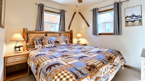 Un dormitorio con una cama grande, cabecera de madera, dos mesitas de noche con lámparas, un ventilador de techo y dos ventanas con cortinas.
