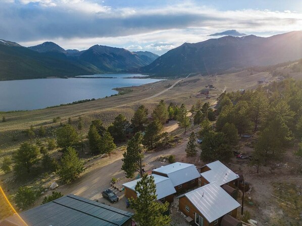 Una cabaña apartada con techo de metal, rodeada por un bosque y un lago, con montañas al fondo.