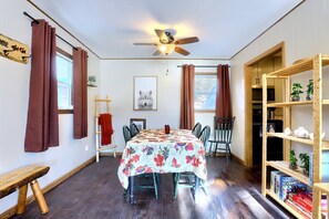 Una habitación con una mesa de comedor, sillas, un ventilador de techo, una estantería y un ventanal con cortinas.