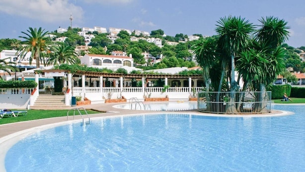 A swimming pool with a surrounding deck, palm trees, and a building with a white facade in the background. - Son Bou Apartments (Alayor)