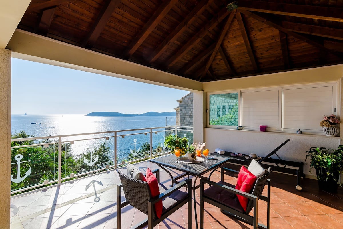 A covered outdoor area with a table and chairs, a view of the sea, and a window with decorative items.