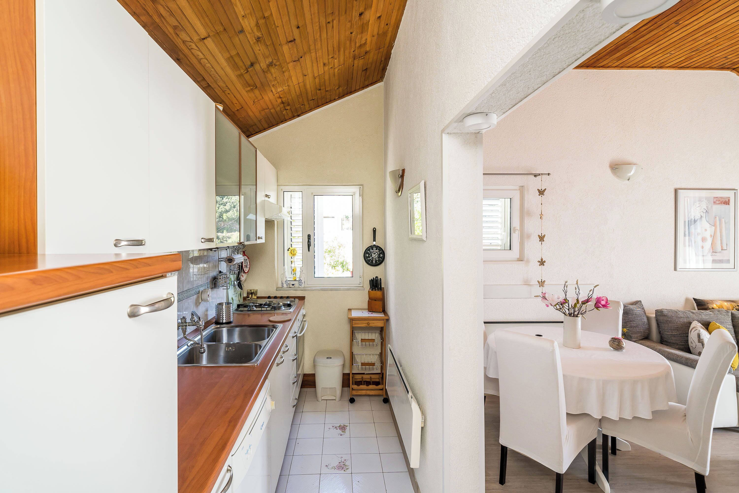 A compact kitchen with wooden cabinets, a sink, and a stove. There is a dining area with a table and chairs, and a sofa in the background.