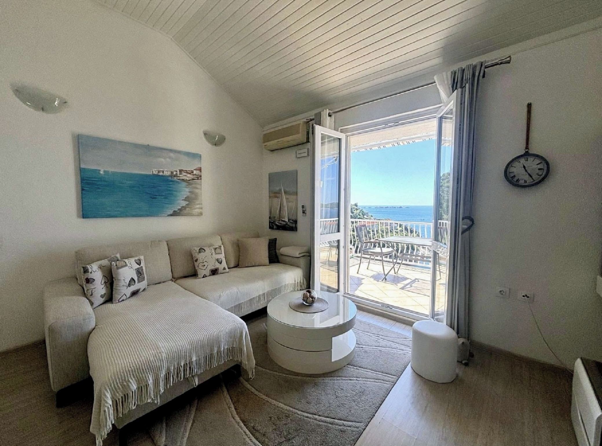 A bedroom with a sofa, a round table, a view of the sea, a clock, and a balcony.
