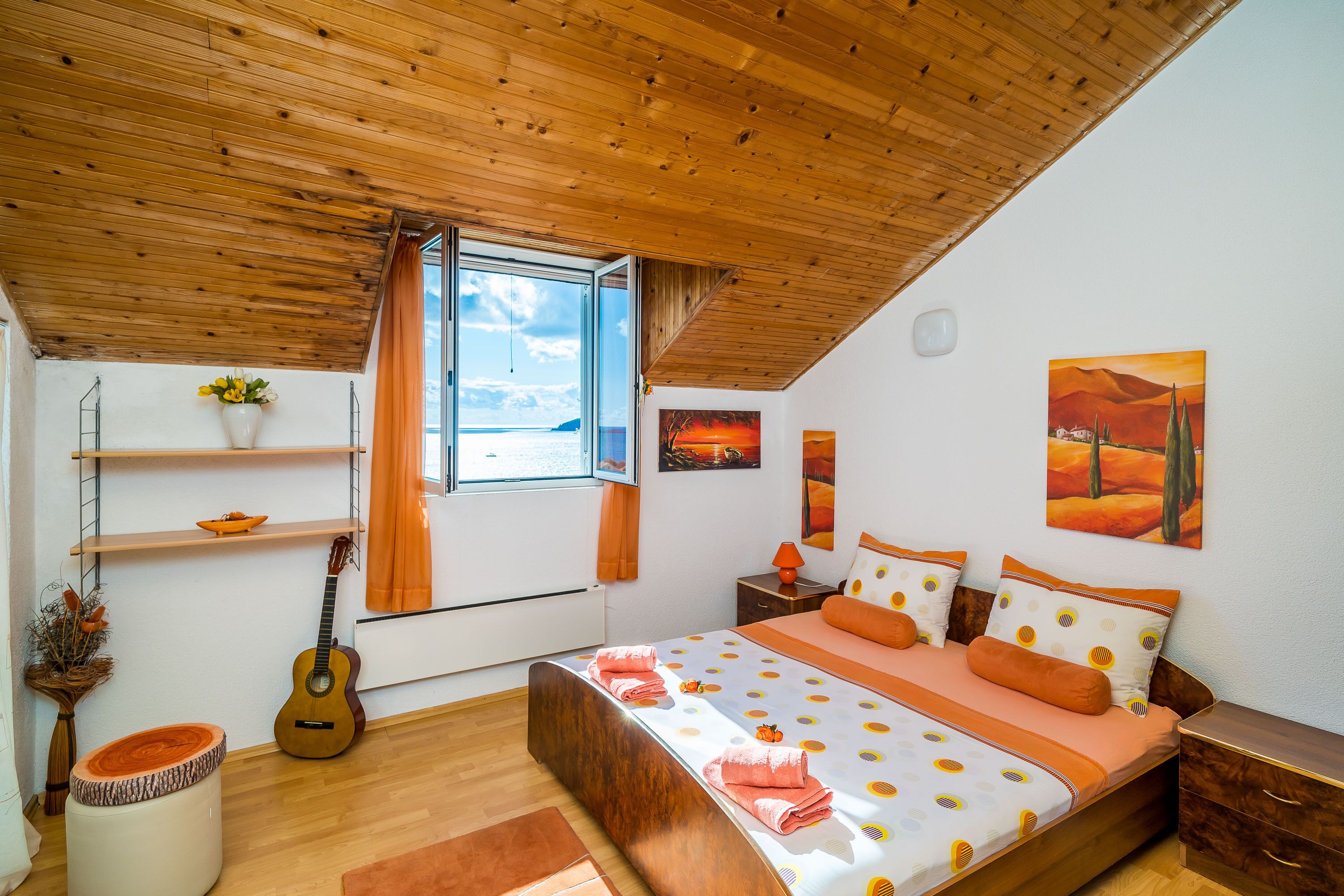 Un dormitorio con techo de madera, cama con ropa de cama naranja, una guitarra, una ventana con cortinas y obras de arte en las paredes.