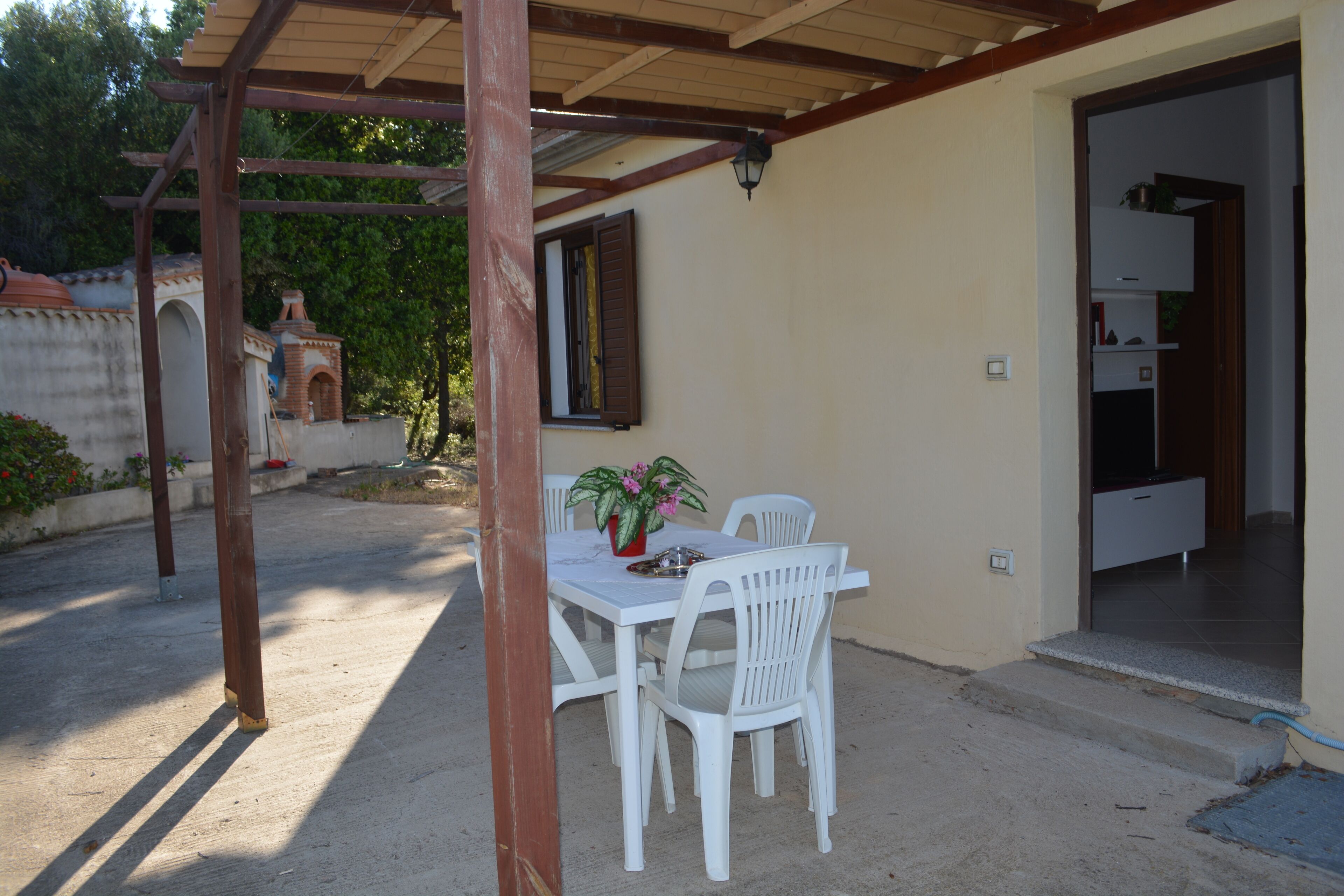 A covered patio with a white table and chairs, a potted plant, and a view of a garden and building in the background.