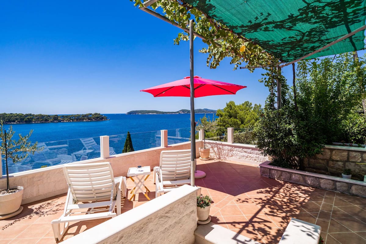 Una terraza con vistas al mar, sillas reclinables blancas, un paraguas rojo y plantas en macetas.