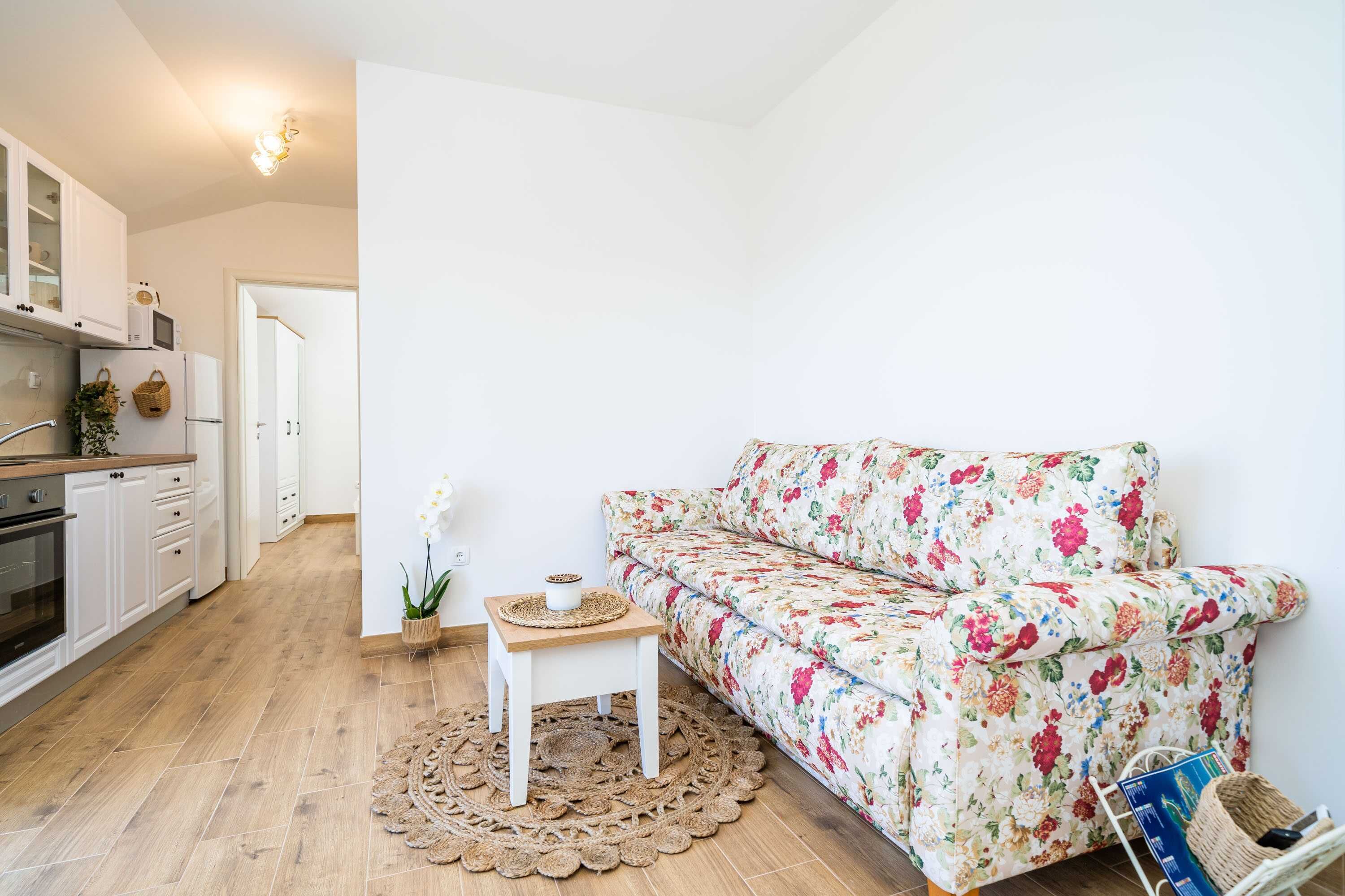 Une cuisine moderne avec des meubles blancs, un four à micro-ondes et un réfrigérateur. Le sol est en bois, il y a un canapé blanc recouvert d’un tissu à motifs floraux, ainsi qu’une petite table sur laquelle sont posés une plante et un bol.