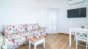 A living room with a floral sofa, a small wooden table, a white chair, a flat-screen TV, and a door with a decorative arch. - Apartments Lovro (Dubrovnik)